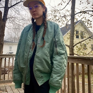 May include: A woman wearing a green bomber jacket with a yellow baseball cap and glasses. She has her hair in two braids. The jacket has a zipper and pockets. The woman is standing on a wooden deck.