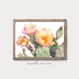 Puede incluir: Una pintura de acuarela de un cactus chumbera con flores rosas y amarillas. El cactus está en un marco de madera y el fondo es blanco. La pintura está firmada "Rainshine Creatives".