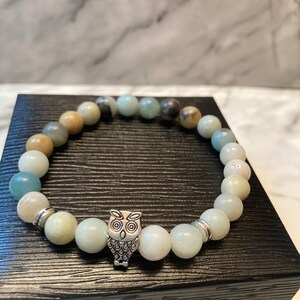 Peut inclure: Un bracelet en perles vertes et brunes avec un charme hibou. Le bracelet est sur une boîte noire.