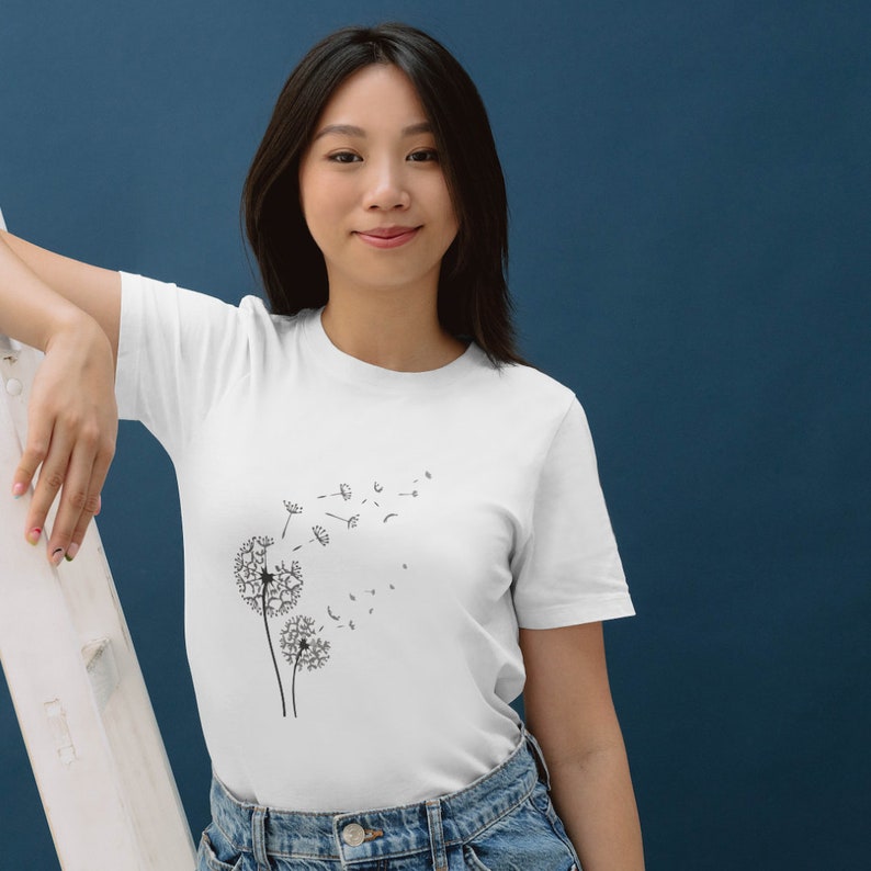May include: A white t-shirt featuring a black dandelion graphic. The dandelion is blowing in the wind and the seeds are scattering.
