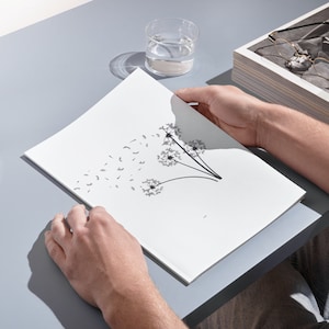 May include: A white booklet featuring a black and white illustration of three dandelions with seeds blowing in the wind. The booklet is on a grey surface.