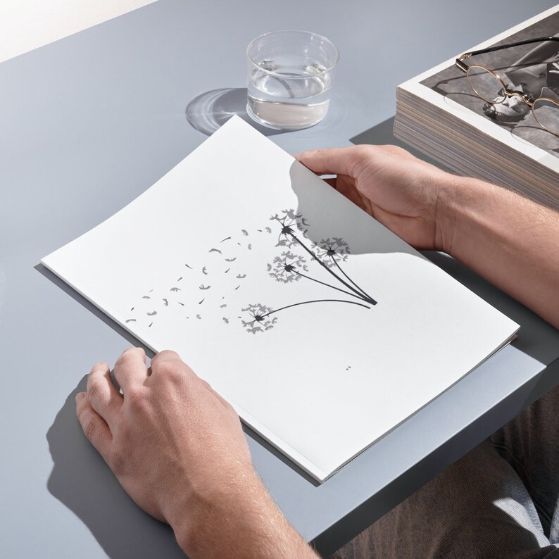 May include: A white booklet featuring a black and white illustration of three dandelions with seeds blowing in the wind. The booklet is on a grey surface.
