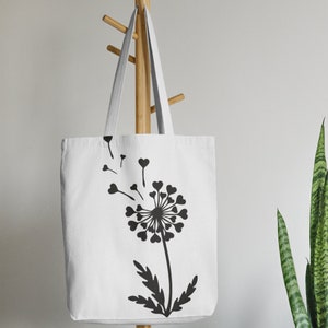 May include: White canvas tote bag featuring a black silhouette of a dandelion with heart-shaped petals and seeds blowing in the wind.