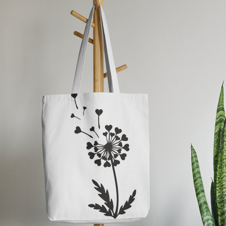 May include: White canvas tote bag featuring a black silhouette of a dandelion with heart-shaped petals and seeds blowing in the wind.