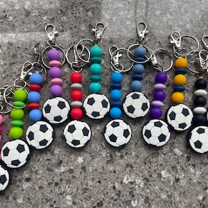 May include: A collection of soccer ball keychains with colorful beaded accents. Each keychain features a black and white soccer ball charm and a variety of bead colors, including pink, green, blue, purple, yellow, black, and red.