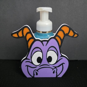 May include: A purple and white soap dispenser featuring a cartoon dragon face with orange horns. The dispenser has a white pump and a blue base. The design is a whimsical, decorative addition to any bathroom.