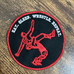 May include: A black and red embroidered patch with a wrestler in a red silhouette. The text around the wrestler reads "Eat, Sleep, Wrestle, Repeat."