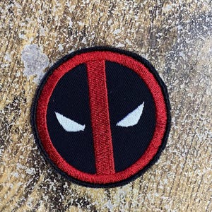 May include: A circular embroidered patch featuring a black background with a thick red border and a vertical red stripe through the center. Two white, almond-shaped eye designs are positioned on either side of the central red stripe.
