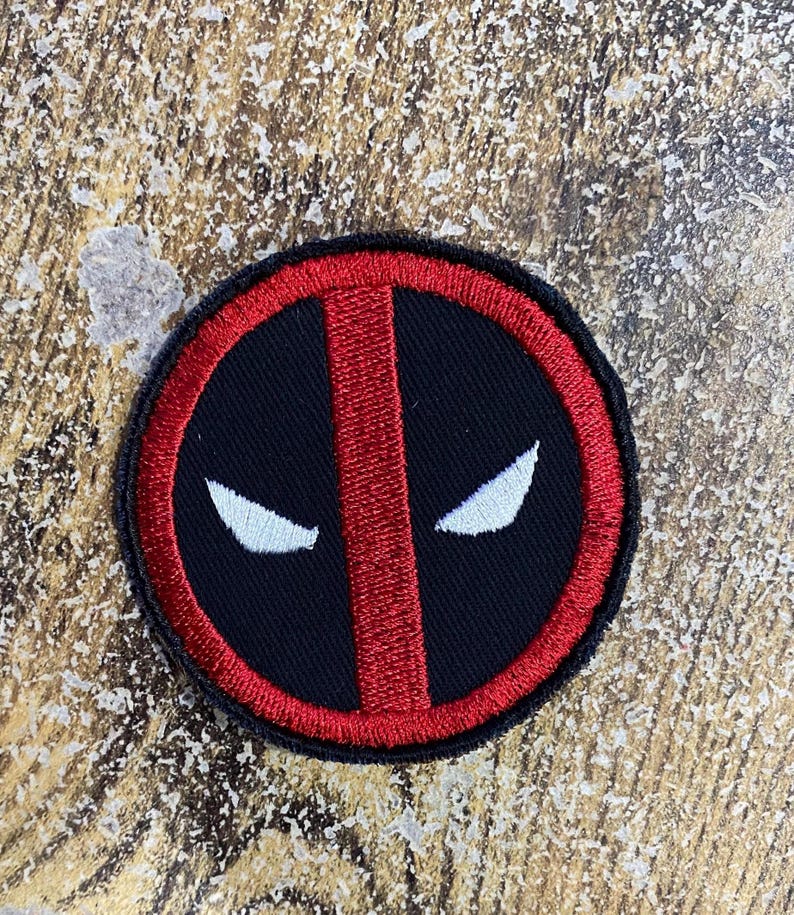 May include: A circular embroidered patch featuring a black background with a thick red border and a vertical red stripe through the center. Two white, almond-shaped eye designs are positioned on either side of the central red stripe.