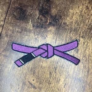 Puede incluir: Cinturón de artes marciales bordado de color morado sobre una superficie de madera marrón.
