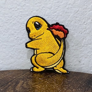 May include: A yellow embroidered patch of a Pokemon character with a flame tail. The character has a blue eye and a black outline.