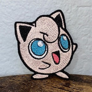 May include: Embroidered patch of a pink Pokemon character with large blue eyes and a red mouth. The character is standing with its arms at its sides.