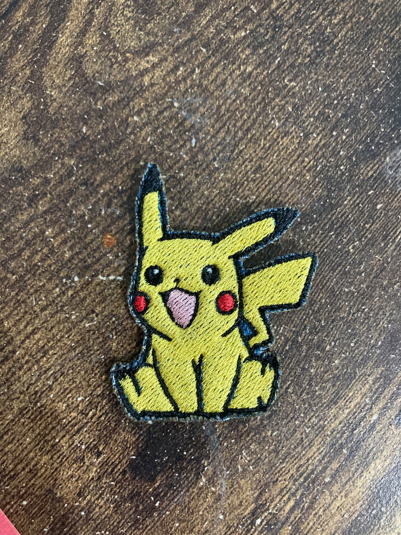 May include: Yellow embroidered patch of Pikachu, a popular cartoon character from the Pokemon franchise. The patch features Pikachu's signature yellow fur, red cheeks, and black eyes.