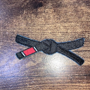 Puede incluir: Parche negro bordado de un cinturón negro atado con una franja roja.