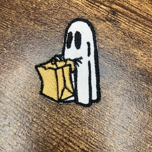 Könnte beinhalten: Ein weißer Geist mit schwarzen Augen, der eine braune Papiertüte hält. Der Geist ist auf einem braunen Stoffhintergrund gestickt.