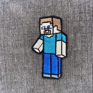 May include: Embroidered patch featuring a pixelated character with a blue shirt, blue pants, and brown hair. The character has a neutral expression and is outlined in black. The patch is likely for a video game enthusiast.