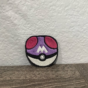 May include: Purple and red embroidered patch of a Pokeball with a white 'M' inside.