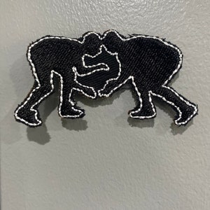 Pode incluir: Etiqueta bordada preta de duas figuras lutando, contornada com fio branco.