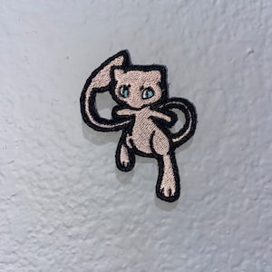 May include: A pink and black embroidered patch of a cartoon pink Pokemon character with blue eyes. The character is standing on its hind legs with its tail curled around its body.