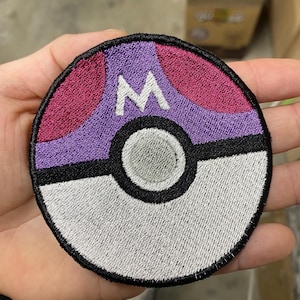 May include: An embroidered patch featuring a purple and pink Pokeball design with a white "M" in the centre.