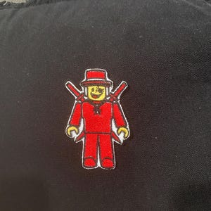 May include: A red embroidered patch featuring a block figure wearing a hat and holding two swords. The figure has yellow hands and a smiling face. The patch is on a black fabric.