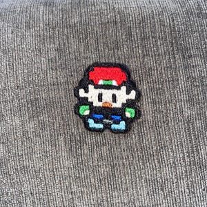 Puede incluir: Un parche pixelado con un personaje con gorra roja, pelo negro y cara blanca. El personaje lleva una camisa negra, pantalones azules y zapatos azules. Acentos verdes en los brazos. El parche está sobre una superficie gris texturizada.