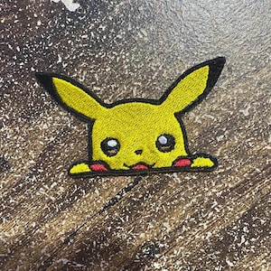 Op de afbeelding: Een geborduurde patch met een geel personage met lange oren met zwarte punten, grote donkere ogen, een kleine neus en rode cirkels op de wangen. Het personage is vanaf de borst afgebeeld en lijkt over een rechte rand heen te kijken.