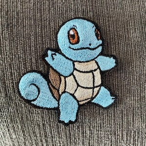 May include: Embroidered patch featuring a light blue cartoon character with a brown shell and orange eyes. The character has outstretched arms and a smiling expression. The patch is outlined in black.