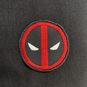 May include: A circular embroidered patch featuring a red border and a vertical red stripe centered on a black background. Two white, almond-shaped eye designs are positioned symmetrically on either side of the central red stripe.