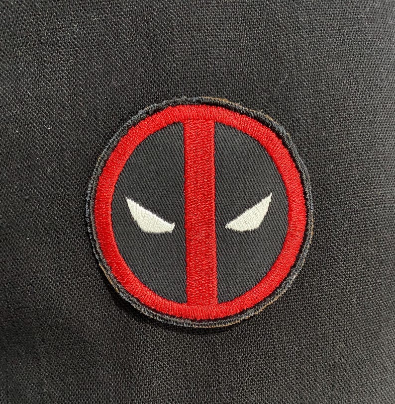May include: A circular embroidered patch featuring a red border and a vertical red stripe centered on a black background. Two white, almond-shaped eye designs are positioned symmetrically on either side of the central red stripe.
