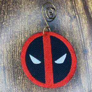 Puede incluir: Adorno de Navidad rojo y negro bordado con el logotipo de Deadpool. El adorno tiene un colgador de alambre dorado.