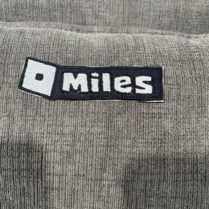 May include: Close-up of a textured, gray fabric with a rectangular patch. The patch is black with the white word "Miles" and a white square with a black dot. The fabric has vertical lines.