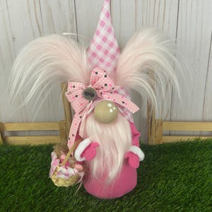 May include: A pink Easter gnome with a long white beard and pink fur hair. The gnome wears a pink gingham hat and a pink bow with black polka dots. It holds a basket with small pink eggs.