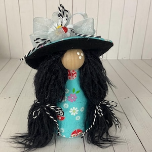 Puede incluir: Una muñeca gnomo caprichosa con cabello negro, un sombrero negro con una cinta blanca y negra, y un vestido floral azul y blanco. El sombrero está decorado con un lazo blanco y una margarita blanca.