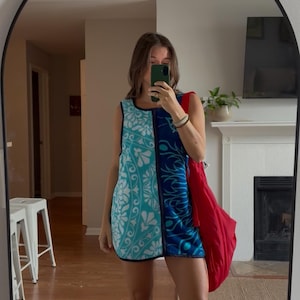 Puede incluir: Un vestido corto sin mangas con un diseño de paneles divididos, que presenta un lado azul claro con un patrón floral blanco y un lado azul oscuro con un motivo de remolinos, junto con un bolso de hombro rojo grande y sandalias beige.