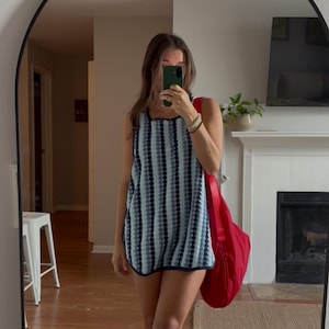 May include: A sleeveless tunic top featuring a vertical striped pattern in shades of light blue, dark blue, and white, accented with a dark blue trim along the neckline and hem, paired with a large, solid red shoulder bag and beige sandals.