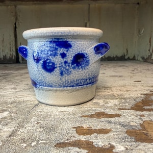May include: A small, cylindrical ceramic crock featuring a speckled off-white base with vibrant blue decorative spots and two side handles. The rim is smooth and rounded, while the lower portion of the vessel remains a solid, unglazed off-white color.