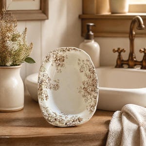 May include: An oval ceramic soap dish featuring a cream-colored base with a brown floral pattern around the rim. The dish has a slightly irregular, organic shape and a smooth, glossy finish, showcasing detailed botanical illustrations.