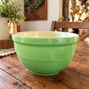 May include: A light green mixing bowl with a smooth, glossy finish and a cream-colored interior. The exterior features a decorative band of vertical ribbed lines and circular patterns around the upper portion of the bowl, with a wide, rounded rim.