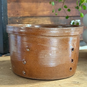 May include: A cylindrical brown ceramic cheese mold featuring a smooth, glazed finish and a thick, reinforced rim. The exterior wall is punctuated by several small, evenly spaced circular drainage holes arranged in a staggered pattern around the body.
