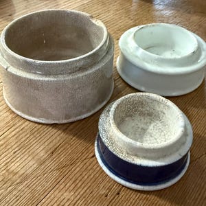 May include: A set of three ceramic containers with a crackled glaze finish. One large beige cylindrical vessel, one white lid with a raised rim, and one smaller lid featuring a dark blue base with a white crackled top section.