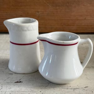 May include: Two white ceramic creamers featuring a thin red horizontal stripe around the upper body. One creamer is taller and cylindrical with a pouring spout, while the other is shorter, bulbous, and includes a side handle and a pouring spout.