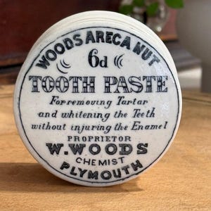 May include: A round white ceramic lid featuring black printed text that reads WOODS ARECA NUT 6d TOOTH PASTE For removing Tartar and whitening the Teeth without injuring the Enamel PROPRIETOR W.WOODS CHEMIST PLYMOUTH with decorative curved lines.