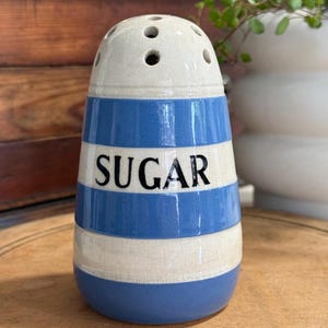 May include: A ceramic sugar shaker featuring a cream-colored base with a crackled glaze finish and horizontal blue stripes. The word SUGAR is printed in black capital letters on the front. The top has multiple holes for dispensing sugar.