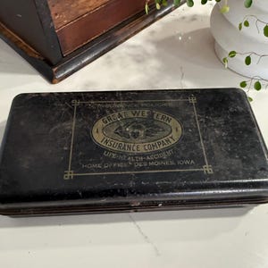 May include: A rectangular black metal document box featuring gold-toned text and graphics. The lid displays the Great Western Insurance Company logo, including the words Life, Health, Accident, and Home Office, Des Moines, Iowa.
