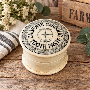 May include: A round, off-white ceramic container with a lid featuring black printed text that reads Calvert's Carbolic Tooth Paste, Trade Mark, and Sixpence, centered around a cross-shaped graphic with a decorative floral border.