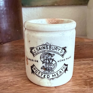 May include: A vintage white ceramic pot with a cylindrical shape and a slightly flared rim. The front features black printed text reading J. SAINSBURY'S SUPERIOR HOME MADE POTTED MEATS, centered around a large decorative letter S with floral details.