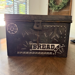 May include: A rectangular dark metal bread box with a hinged lid and a central latch. The front features the word BREAD in white lettering, surrounded by painted wheat stalks, a circular floral motif, and a geometric border along the bottom edge.