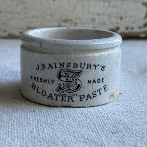 May include: A small, cylindrical white ceramic pot with a crackled glaze finish. The front features black printed text reading J.SAINSBURY'S FRESHLY MADE BLOATER PASTE, centered around a large letter S logo containing the word AINS and a leaf design.