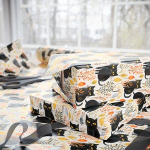 May include: Two gift boxes wrapped in patterned paper featuring black cats, autumn leaves, and foliage. A roll of matching wrapping paper, scissors, and a black ribbon are also visible, suggesting a theme of gift wrapping.